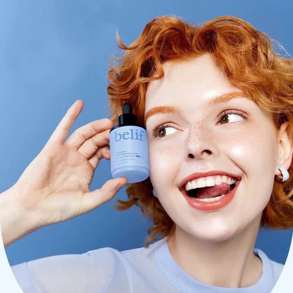 Femme aux cheveux roux tenant un flacon de sérum Belif Super Drops sur fond bleu.