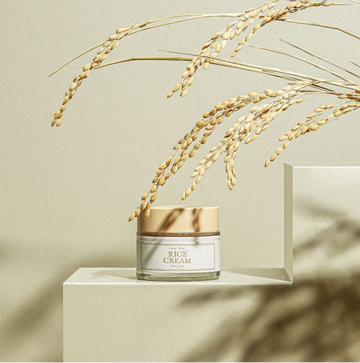 Pot de crème avec couvercle beige sur une plateforme blanche, avec des épis de riz en arrière-plan.