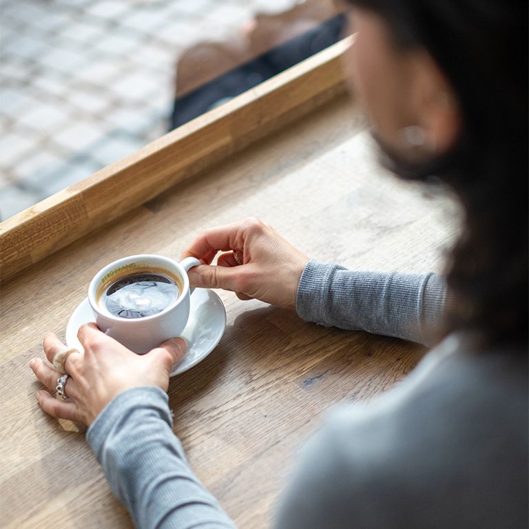 Persoon houdt een kopje koffie op een schotel. Houten oppervlak. Raam op de achtergrond.