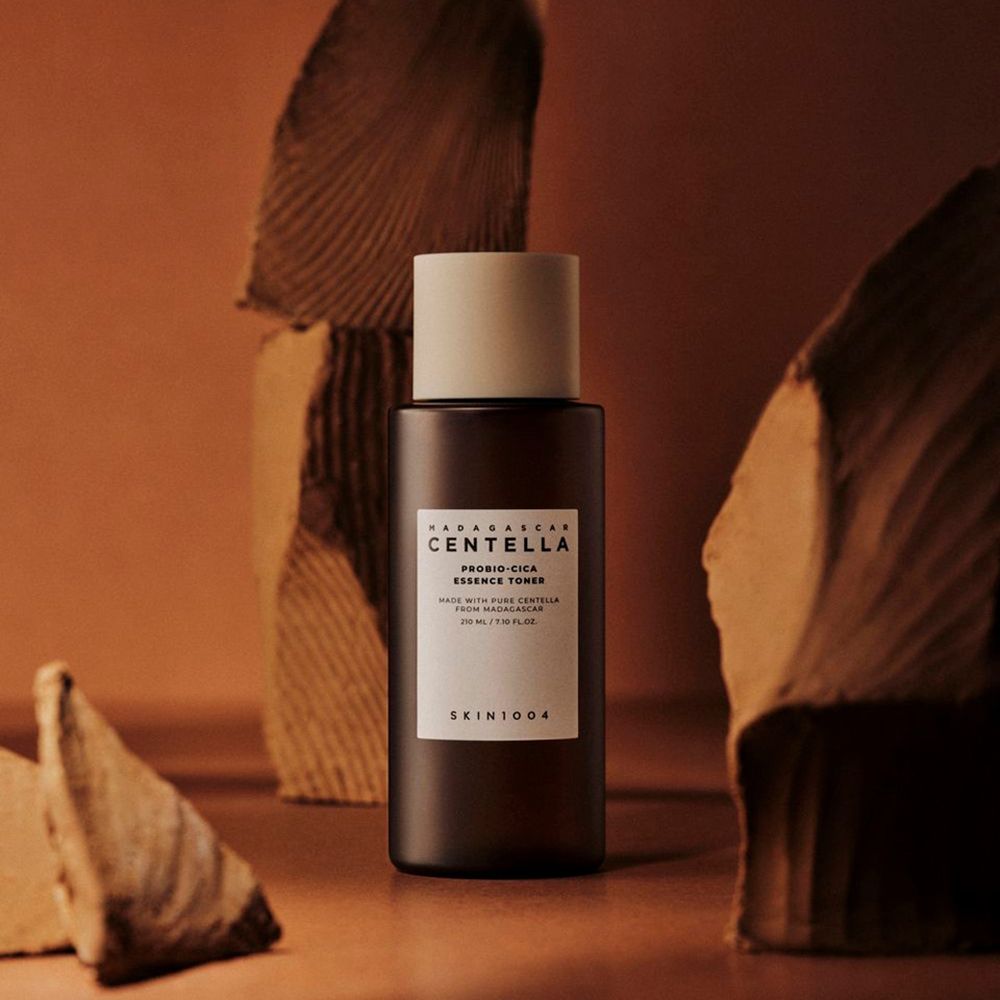 Bruine fles met witte dop. Label met tekst: CENTELLA, PROBIO-CICA ESSENCE TONER, SKIN1004. Op bruine achtergrond.