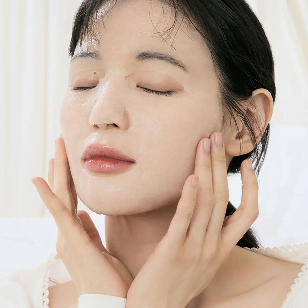 Vrouw met gesloten ogen draagt een sheetmasker, handen op het gezicht.