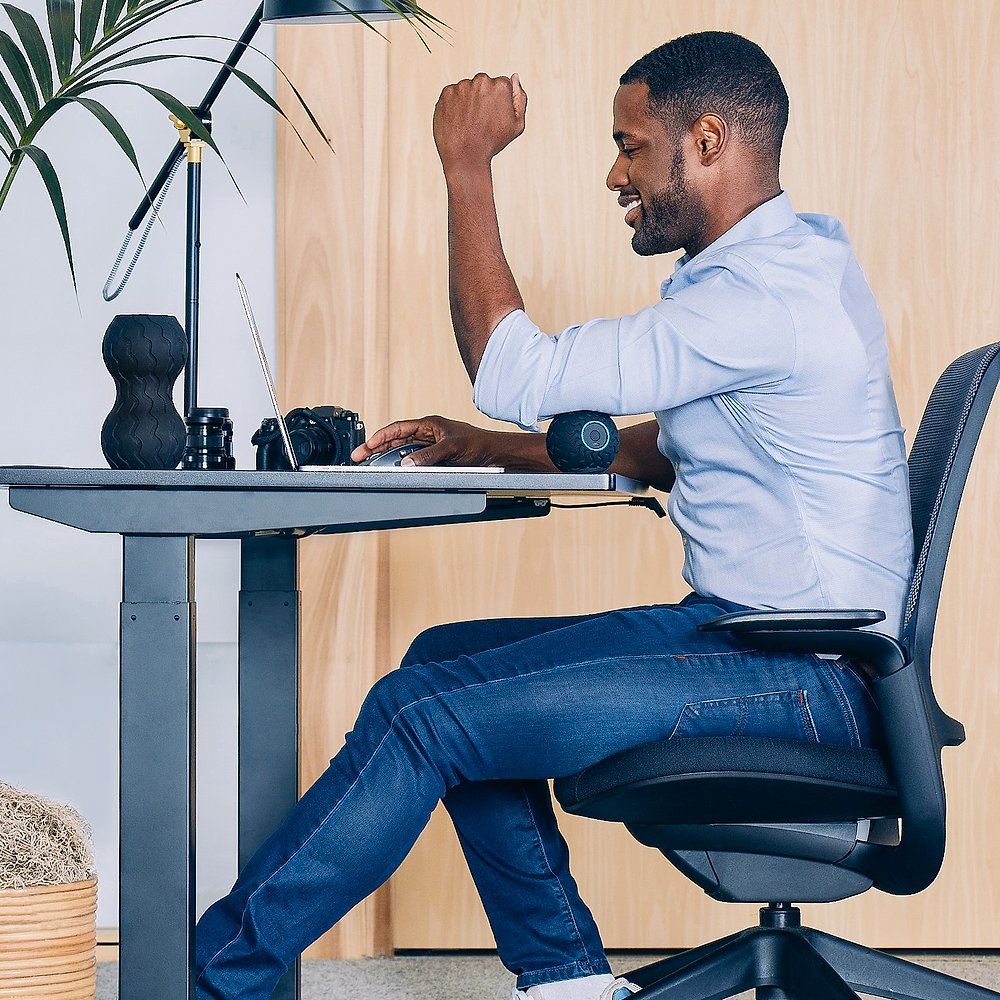 Homme assis à un bureau, la balle de massage sur le bras. Il lève le poing. Ordinateur portable et lampe visibles.
