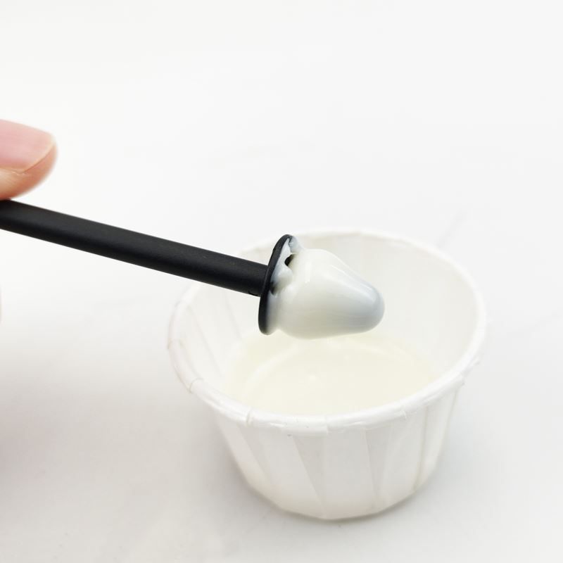 Un applicateur avec de la cire blanche est plongé dans une petite coupelle en papier contenant de la cire liquide.