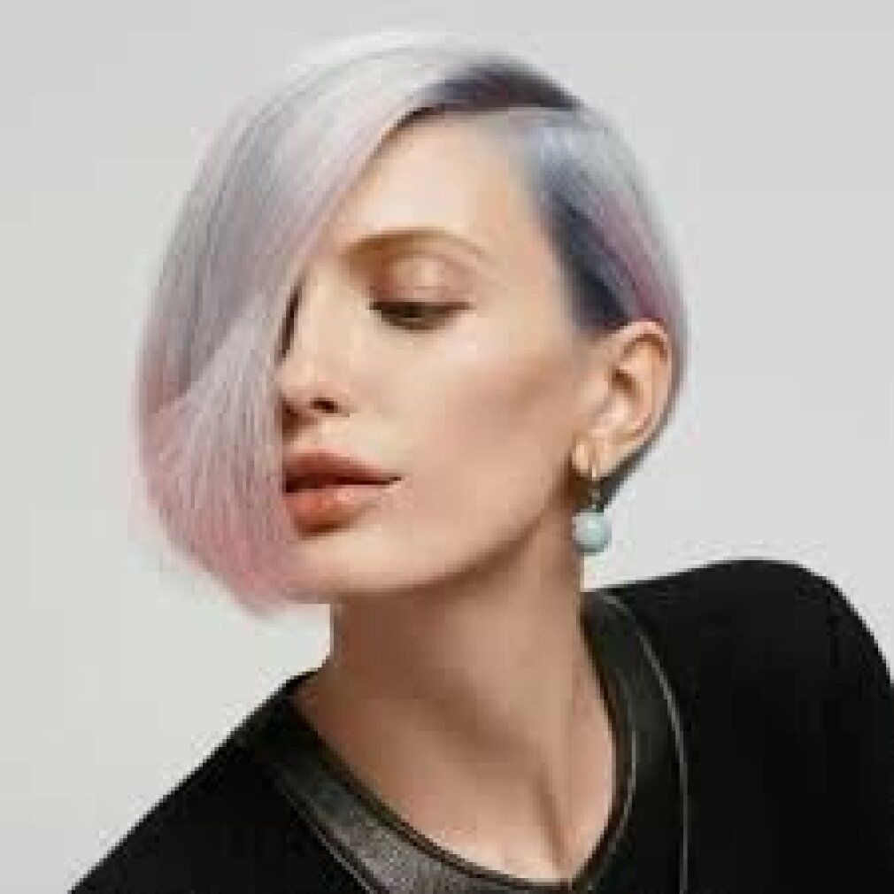 Portrait d'une femme aux cheveux blonds teints en rose et violet. Elle porte un pull noir et des boucles d'oreilles.