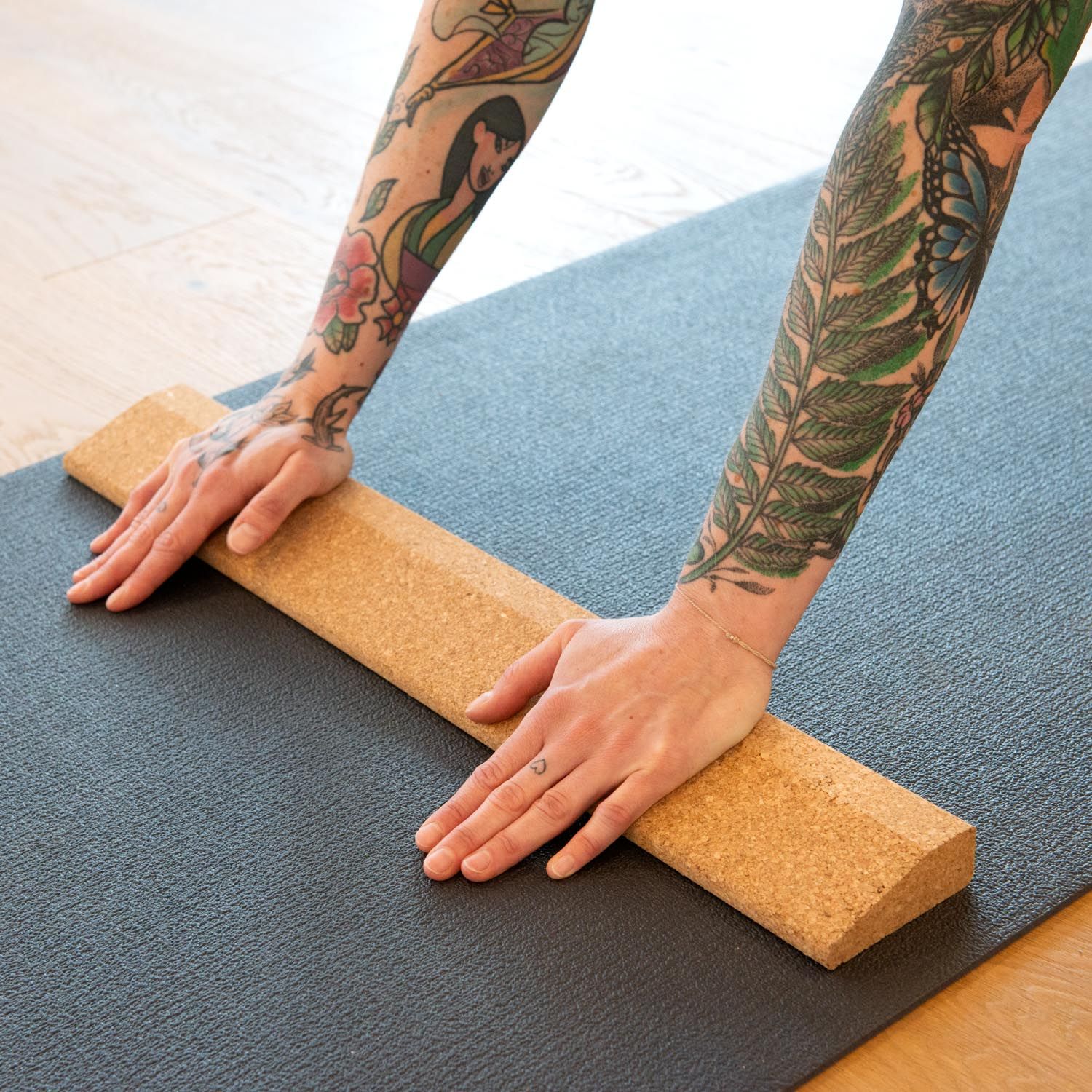 Personne tenant la planche de handstand en liège sur un tapis de yoga. Bras tatoués. Couleur marron.