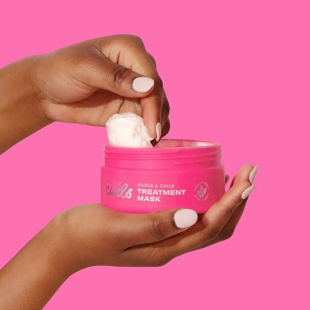 Handen houden een roze pot vast. Een vinger neemt product. Opschrift: "CURLS & COILS TREATMENT MASK".