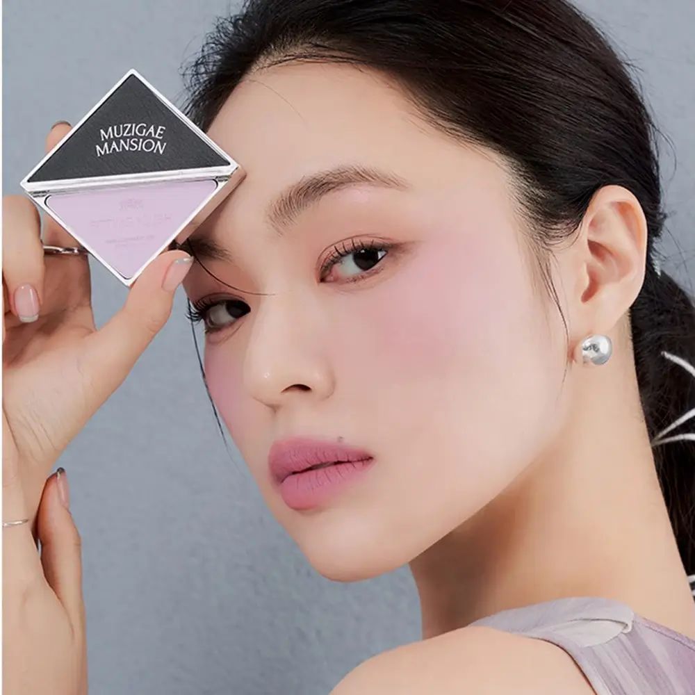 Vrouw houdt vierkante rougeverpakking vast. Zwart deksel met MUZIGAE MANSION. Roze rouge. Vrouw draagt rouge op wangen.