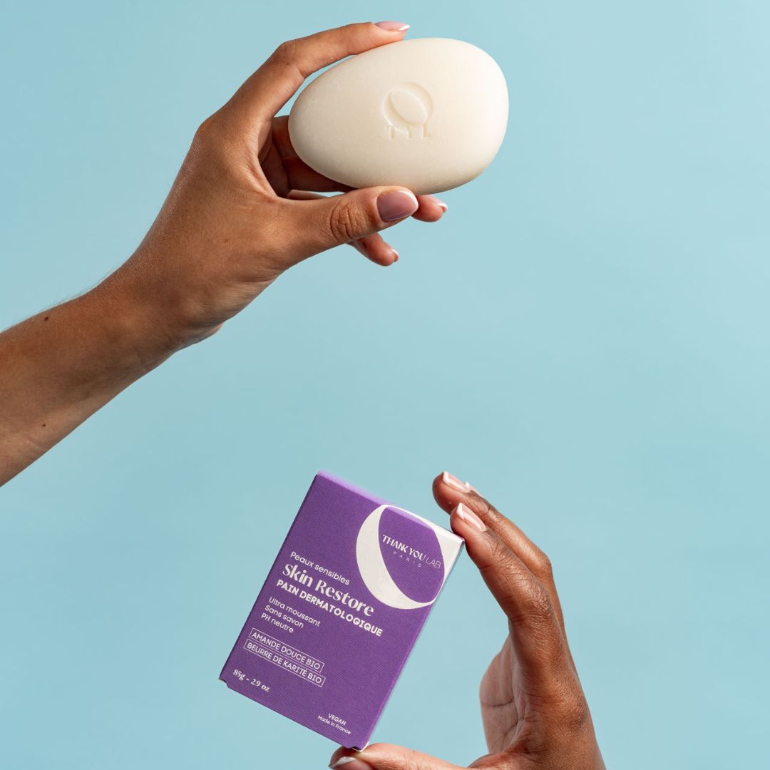 Mains tenant un pain de savon ovale et une boîte violette. La boîte indique "Skin Restore".