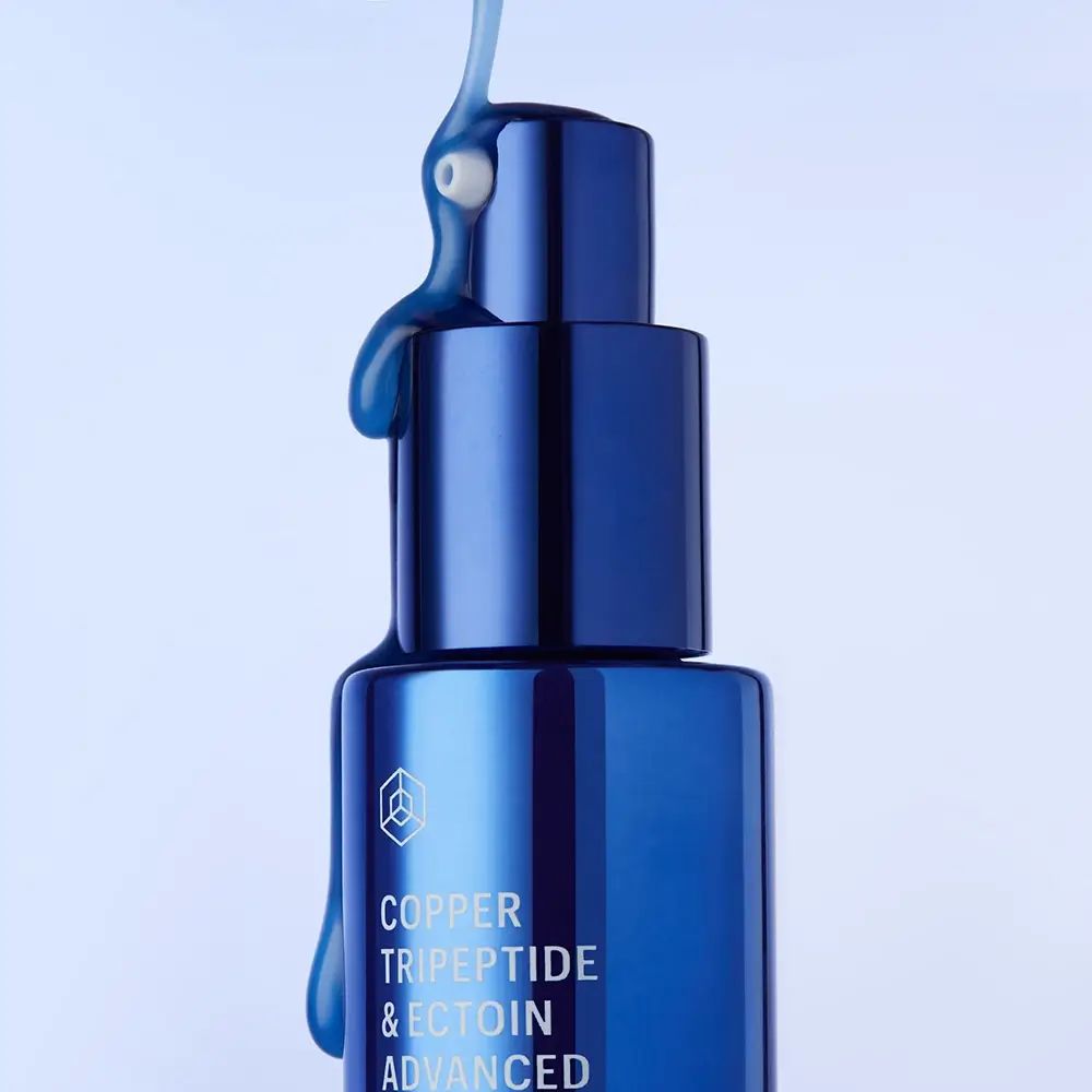 Blauwe metalen fles met serum. Tekst: Copper Tripeptide & Ectoin Advanced Repair Serum. Vloeistof druppelt.