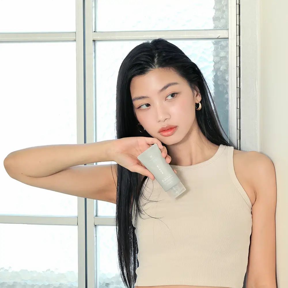 Jonge vrouw houdt een grijze tube vast. Ze draagt een beige top. Achtergrond: raam en witte muur.