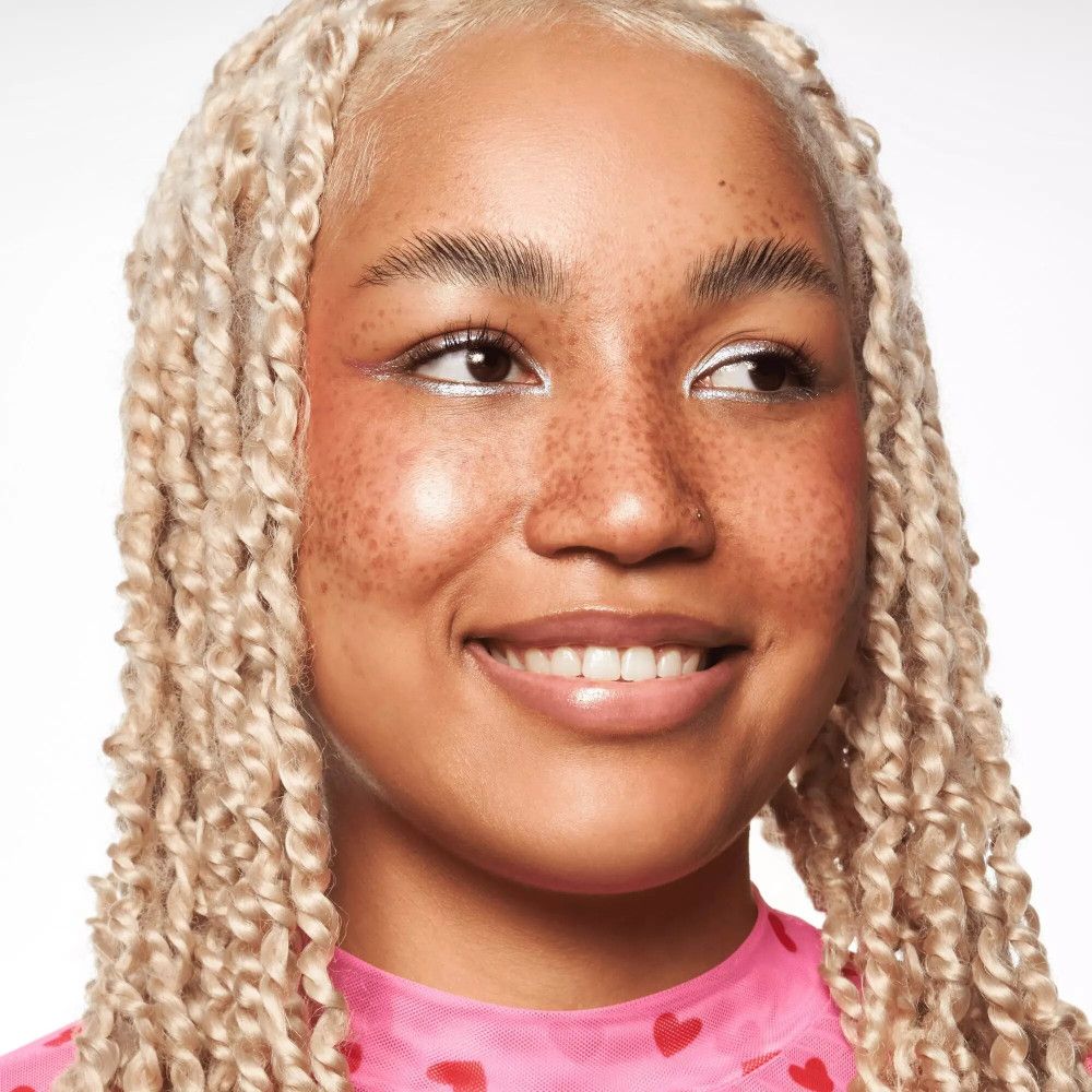 Portrait d'une femme aux tresses blondes. Elle porte un haut rose. Son visage est maquillé avec du fond de teint.