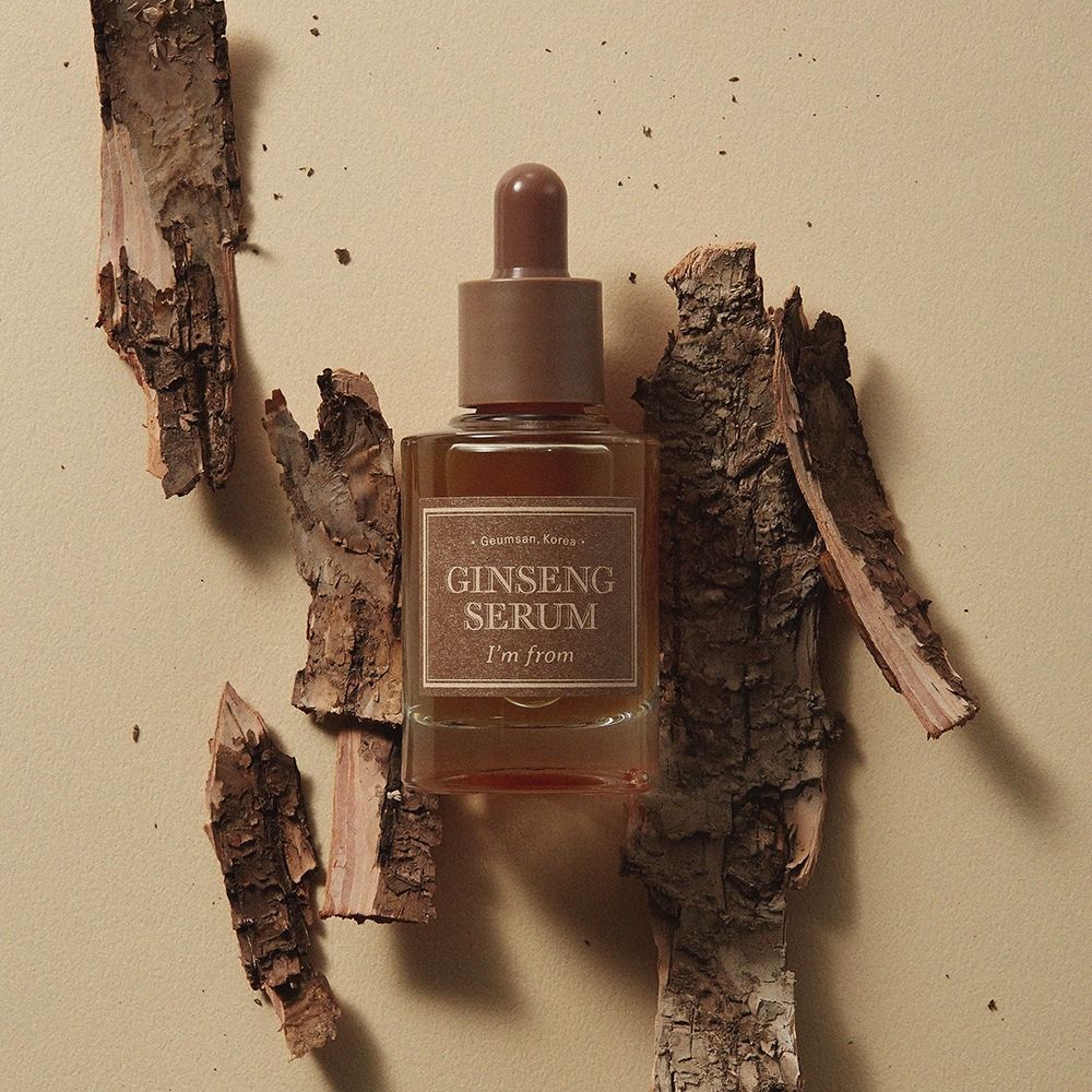 Flacon de sérum avec bouchon marron et étiquette. Entouré de morceaux de bois. L'étiquette indique "Ginseng Serum" et "I'm from".