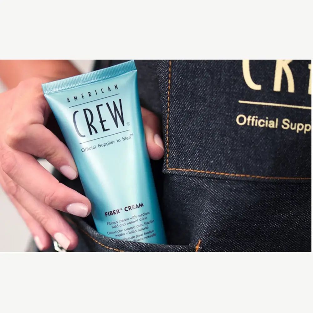 Lichtblauwe tube in een hand. Opschrift: American Crew, Fiber Cream. Op jeans achtergrond.