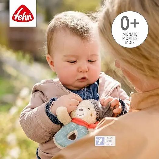 Baby houdt pluche beer rammelaar vast. Fehn-logo linksboven. Leeftijdsadvies 0+ maanden. Rammelaar-icoon.