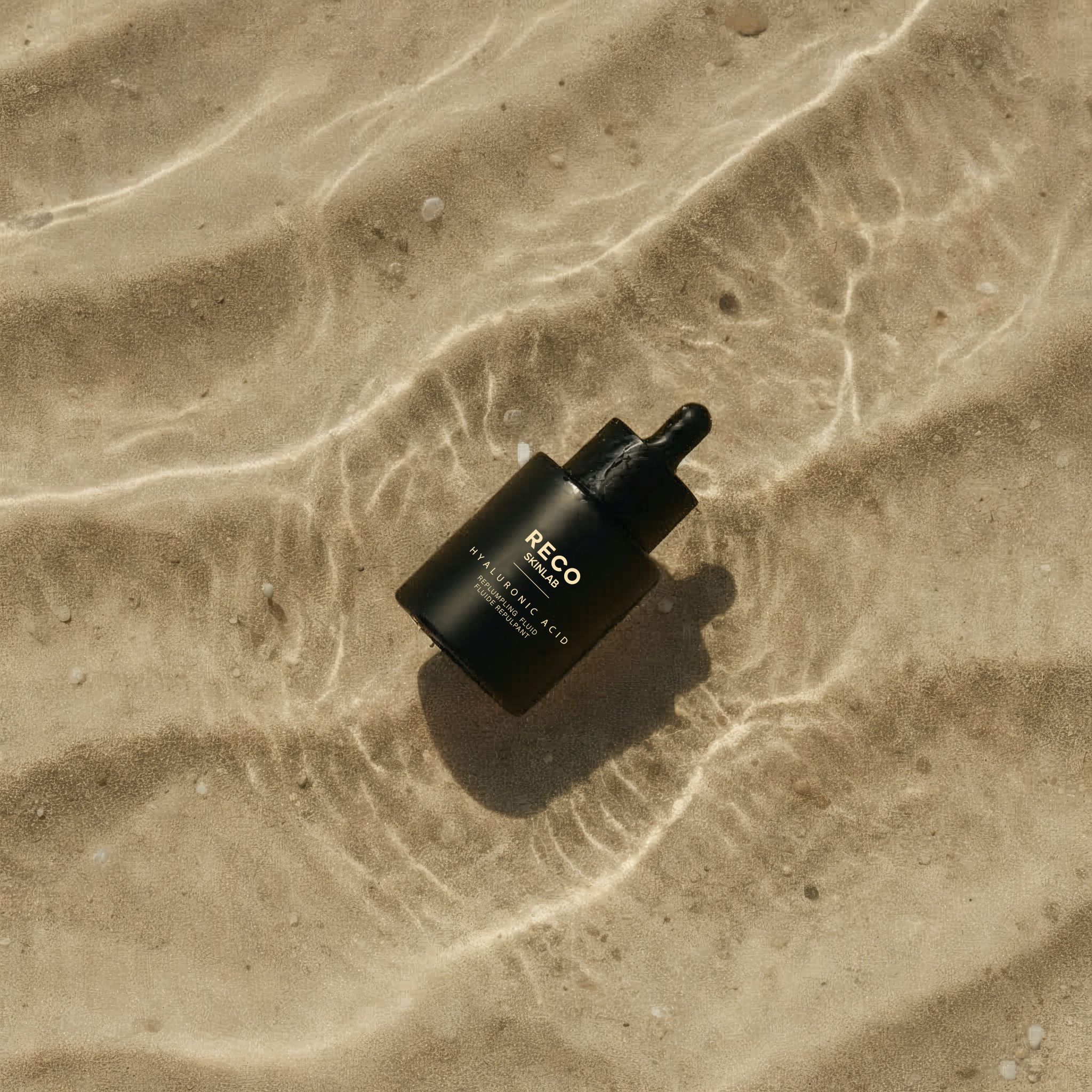 Zwarte fles met pipet in water. Opschrift: RECO SKINLAB, HYALURONIC ACID, REPLUMPING FLUID.