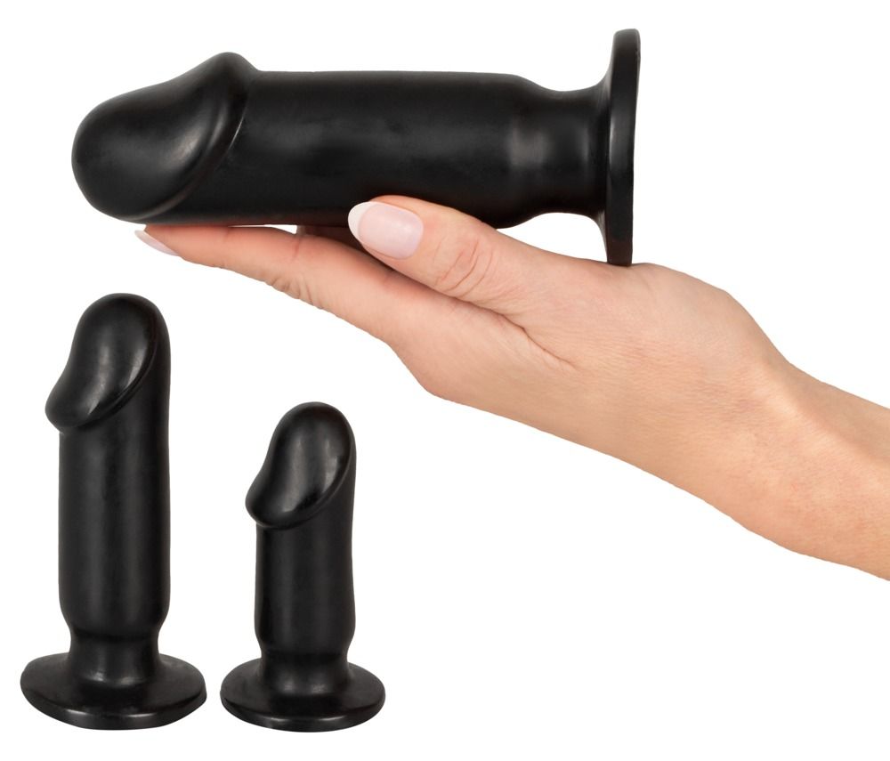 Trois plugs anaux noirs. Un plug est tenu par une main. Les deux autres sont à côté. Forme cylindrique avec pointe arrondie.