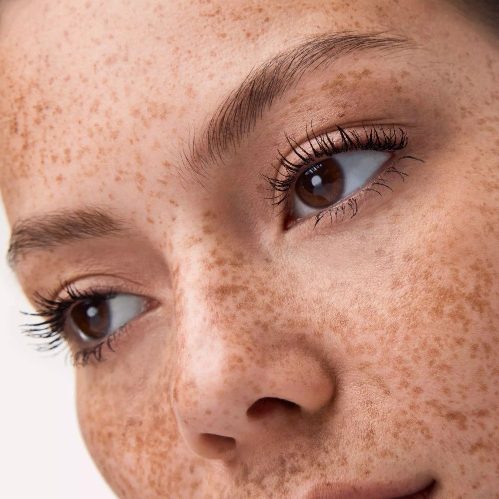 Gros plan d'un visage avec des cils maquillés. La peau est couverte de taches de rousseur.