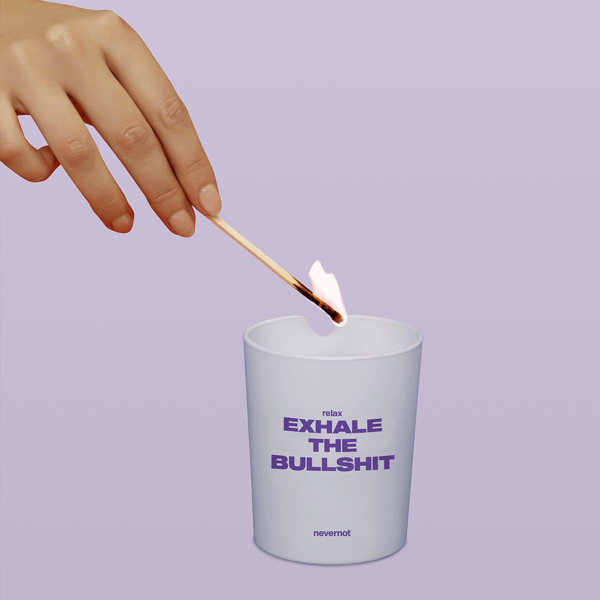 Hand steekt kaars aan. Kaarsbeker met opschrift "relax EXHALE THE BULLSHIT". Merk: nevernot. Achtergrond: lichtpaars.