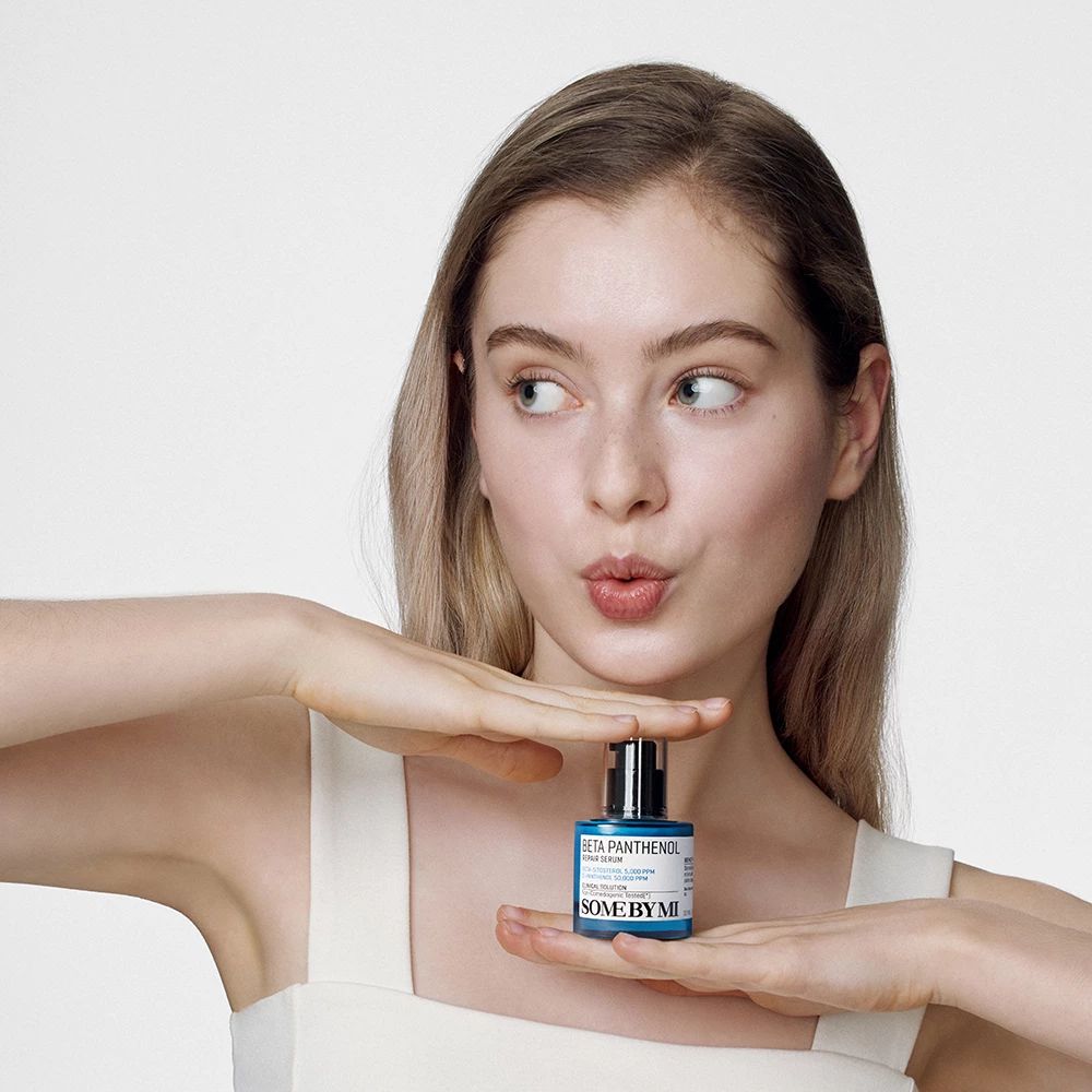 Jeune femme tenant un flacon bleu. Inscription: Beta Panthenol Repair Serum, SOME BY MI. Fond blanc.