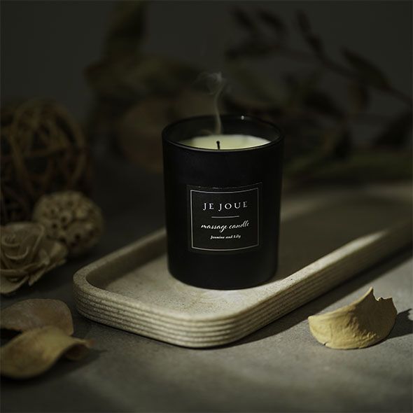 Bougie noire fumante sur un plateau. Inscription : Je Joue, massage candle, Jasmine Lily. Décoration en arrière-plan.