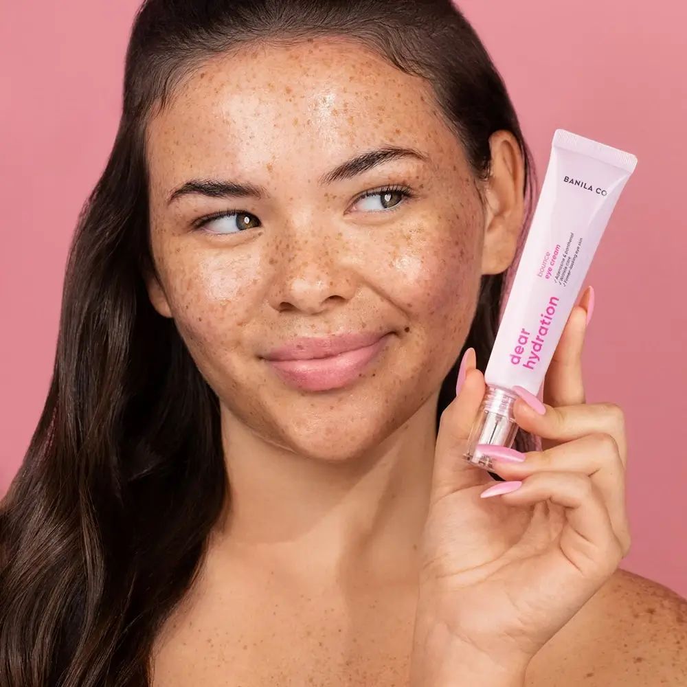 Femme tenant un tube rose de crème pour les yeux. Le tube porte l'inscription "BANILA CO Dear Hydration".
