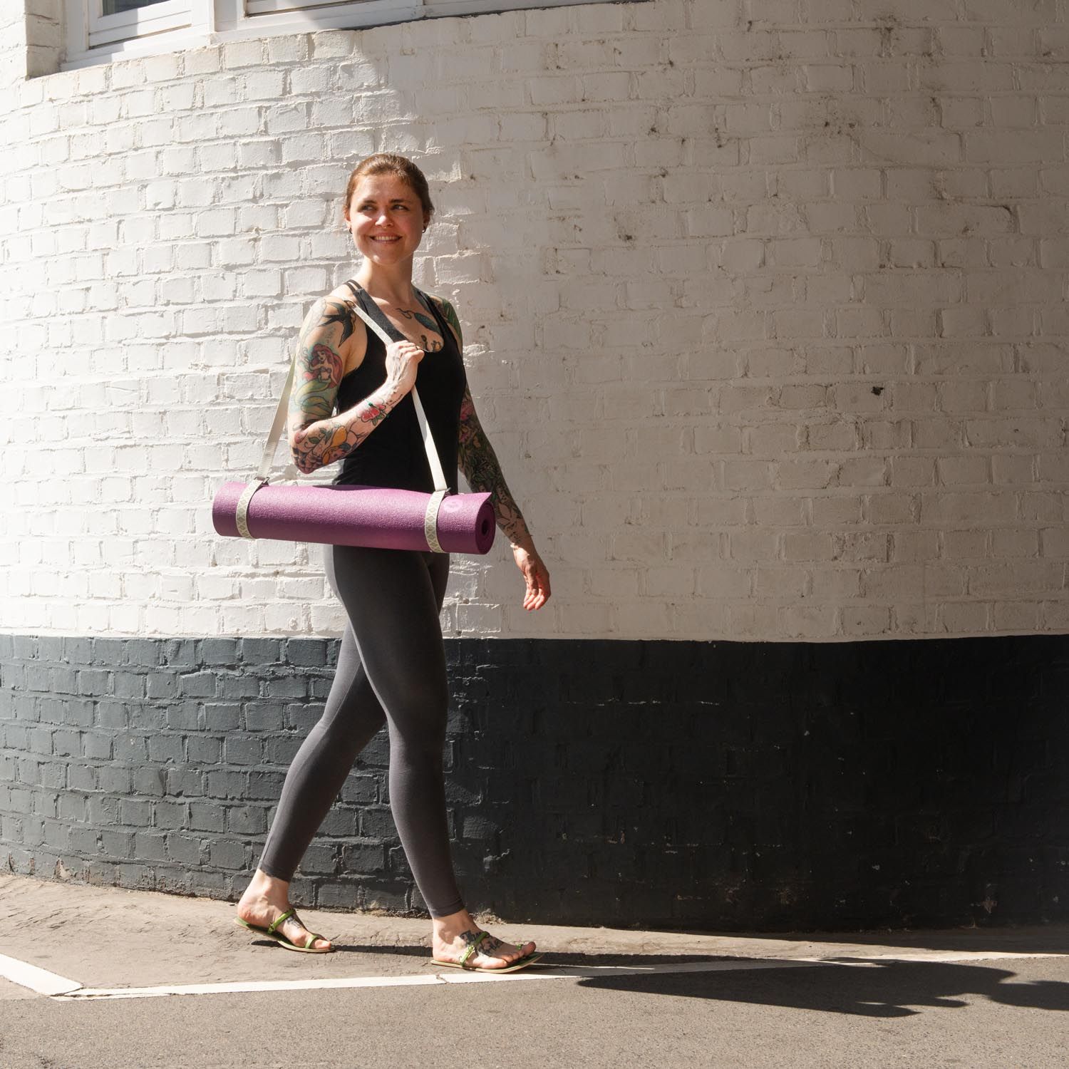 Persoon draagt een paarse yogamat met een lichte draagriem. Loopt langs een witte muur.
