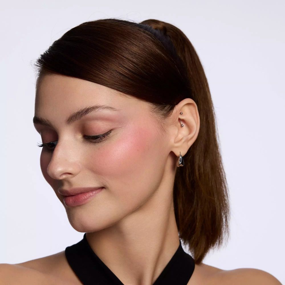 Portrait d'une femme avec du blush rose sur les joues. Elle porte un haut noir et a une queue de cheval. Boucle d'oreille.