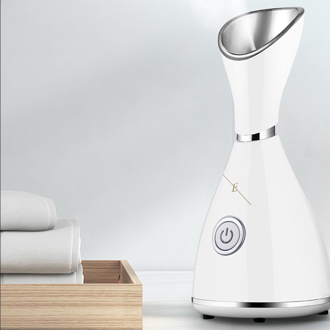 Vaporisateur facial blanc avec tête de vapeur argentée. Sur une table, à côté de serviettes et d'un bac en bois.