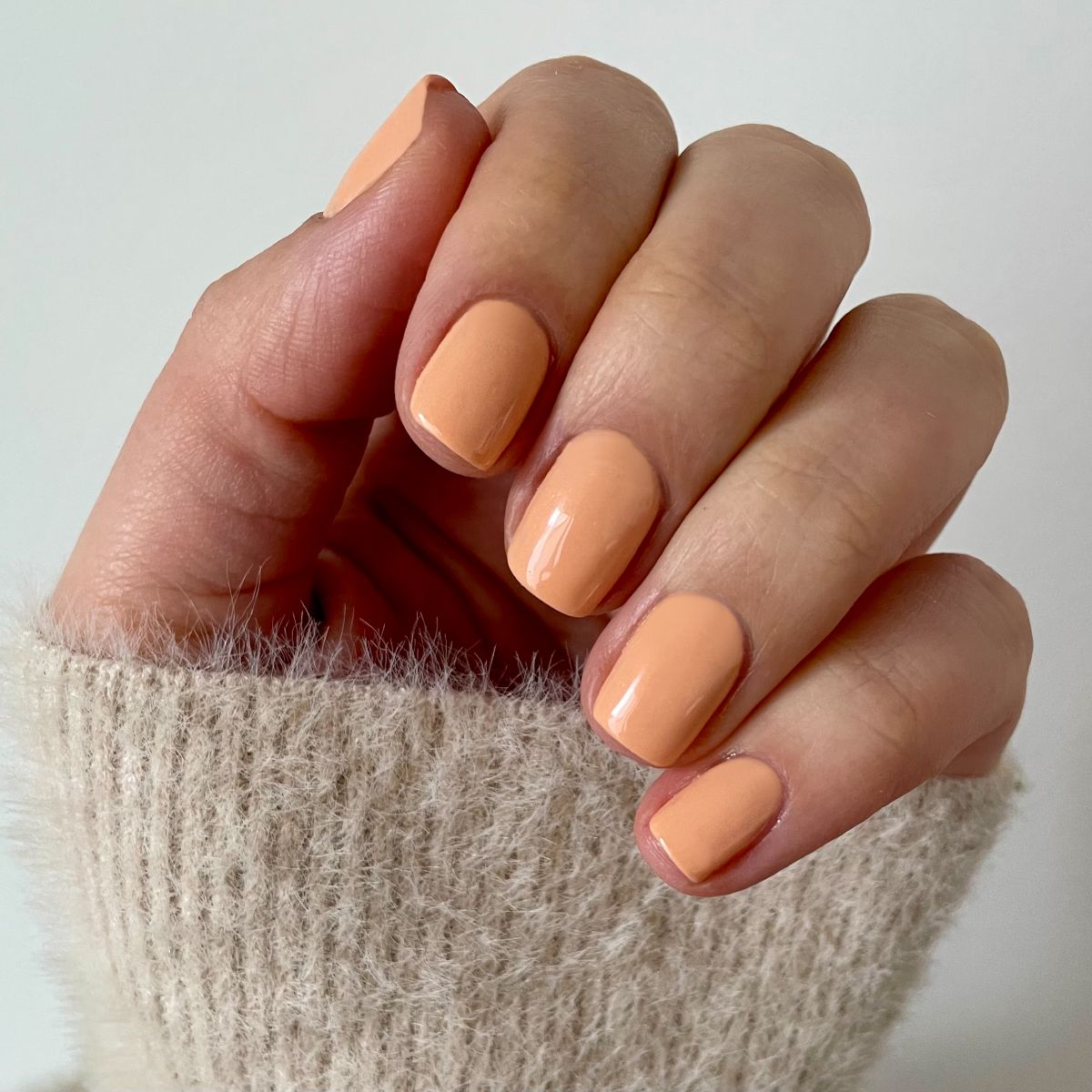 Main avec ongles vernis couleur pêche pastel. Tous les ongles sont courts et vernis. La personne porte un pull beige.