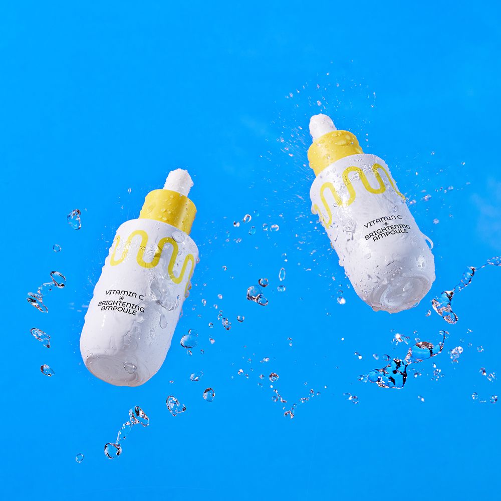 Deux flacons compte-gouttes blancs avec bouchons jaunes, entourés d'eau. Contient Vitamin C Brightening Ampoule.