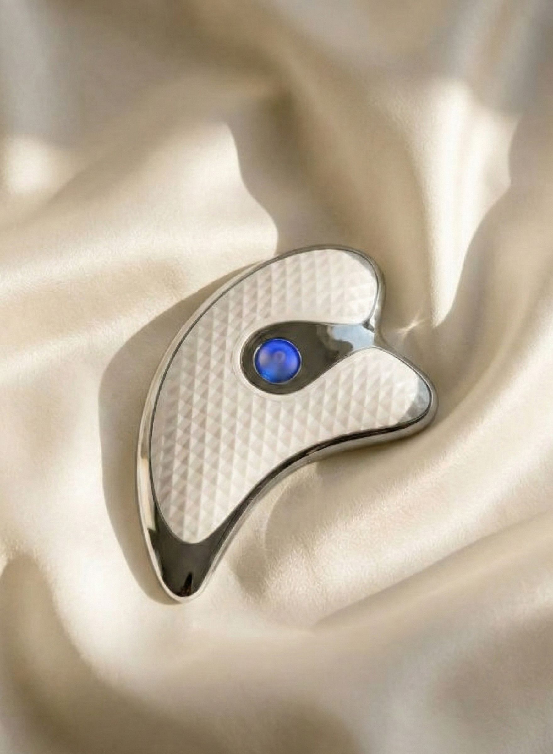 Gua Sha argenté avec un bouton bleu sur un tissu soyeux.