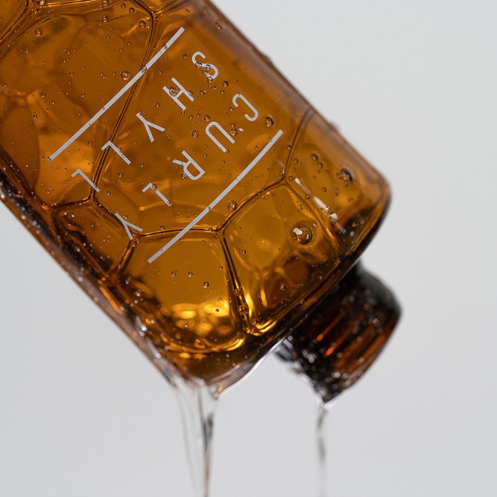 Flacon de shampooing brun, liquide qui coule. Inscription : CURLY SHYLL. Bouteille avec structure en nid d'abeille. Liquide qui coule.