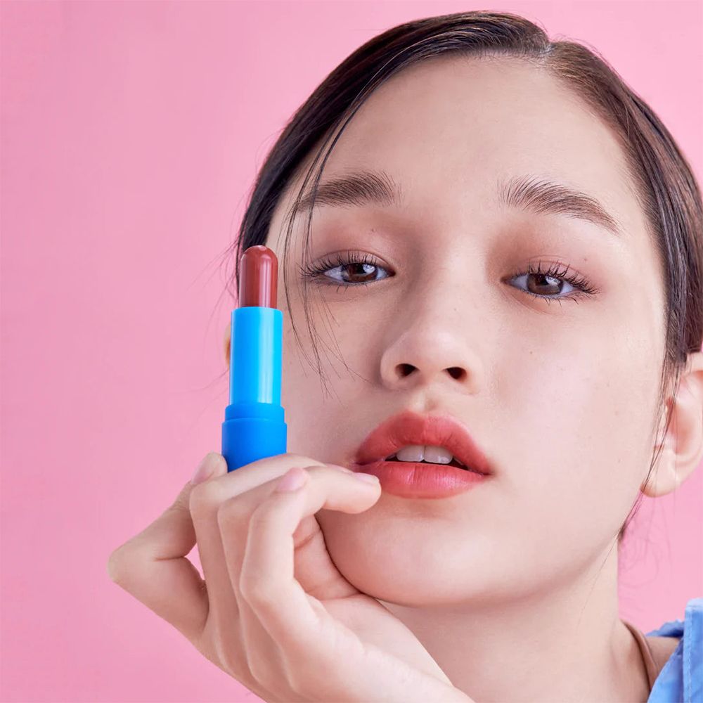 Vrouw houdt lippenbalsem voor haar lippen. Blauwe stift, rode lippenbalsem. Roze achtergrond.