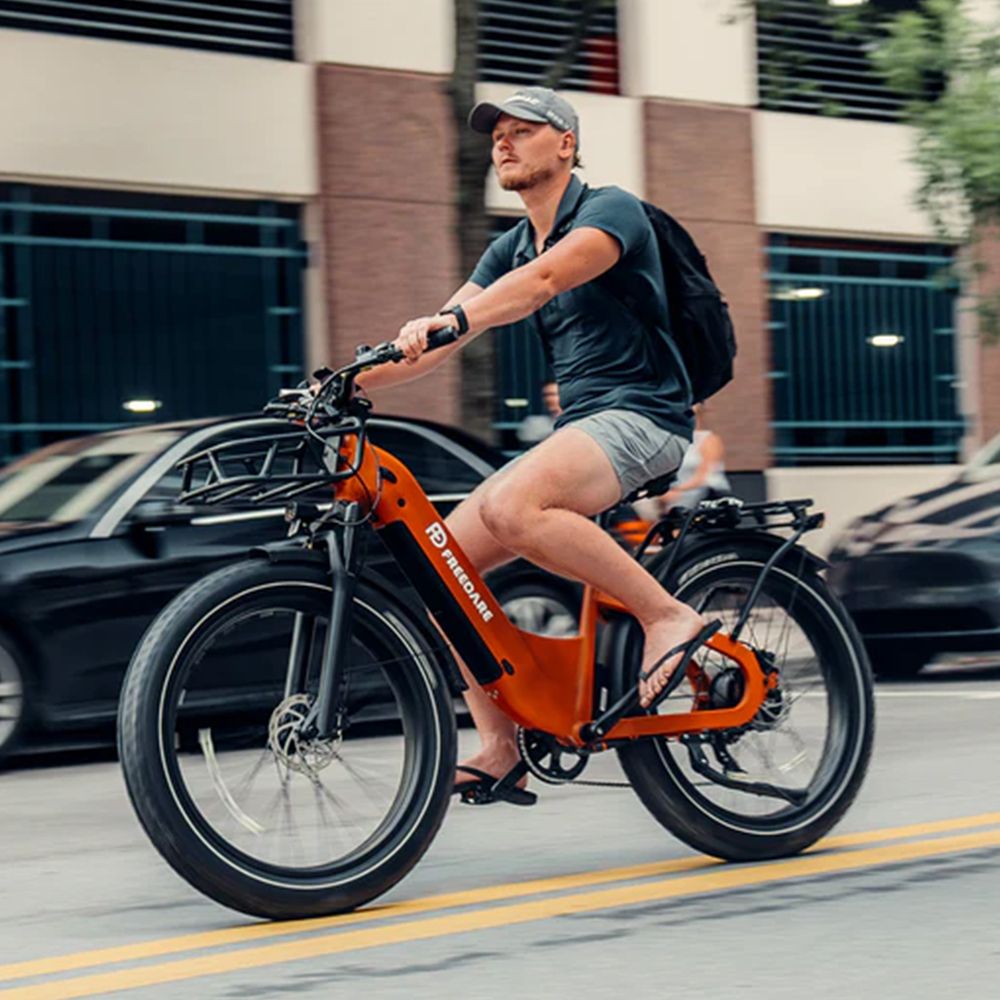 Homme faisant du vélo électrique orange sur une route. Marque FREEDARE. Pneus noirs, porte-bagages.