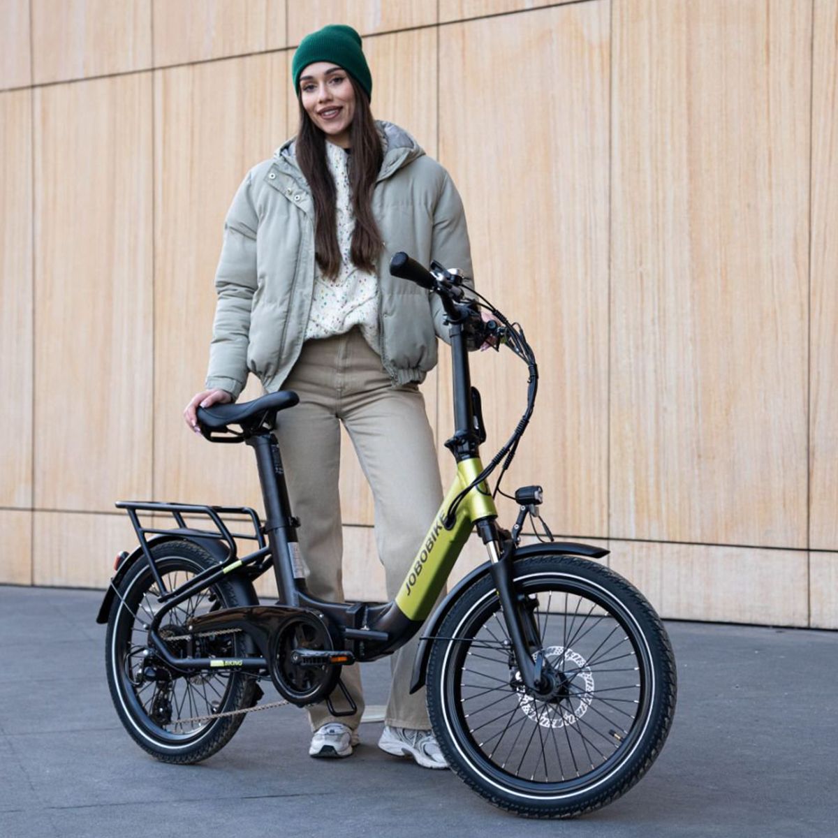 Vélo électrique pliable vert et noir. Logo JOBOBIKE. Porte-bagages, garde-boue. Personne à côté du vélo.