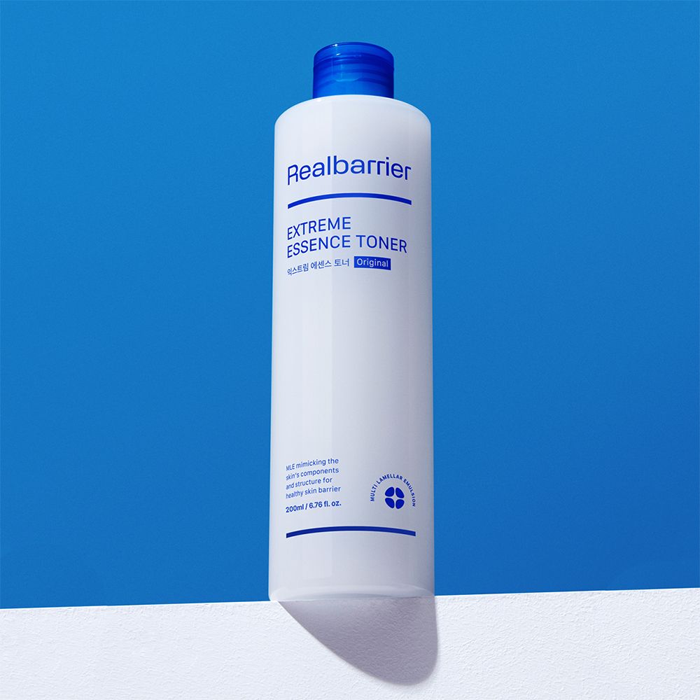 Witte fles met blauwe dop. Opschrift: Realbarrier, Extreme Essence Toner. Fles staat op wit oppervlak.