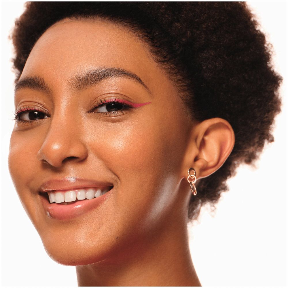 Portrait d'une femme avec un eye-liner rose. Elle porte des boucles d'oreilles et sourit.