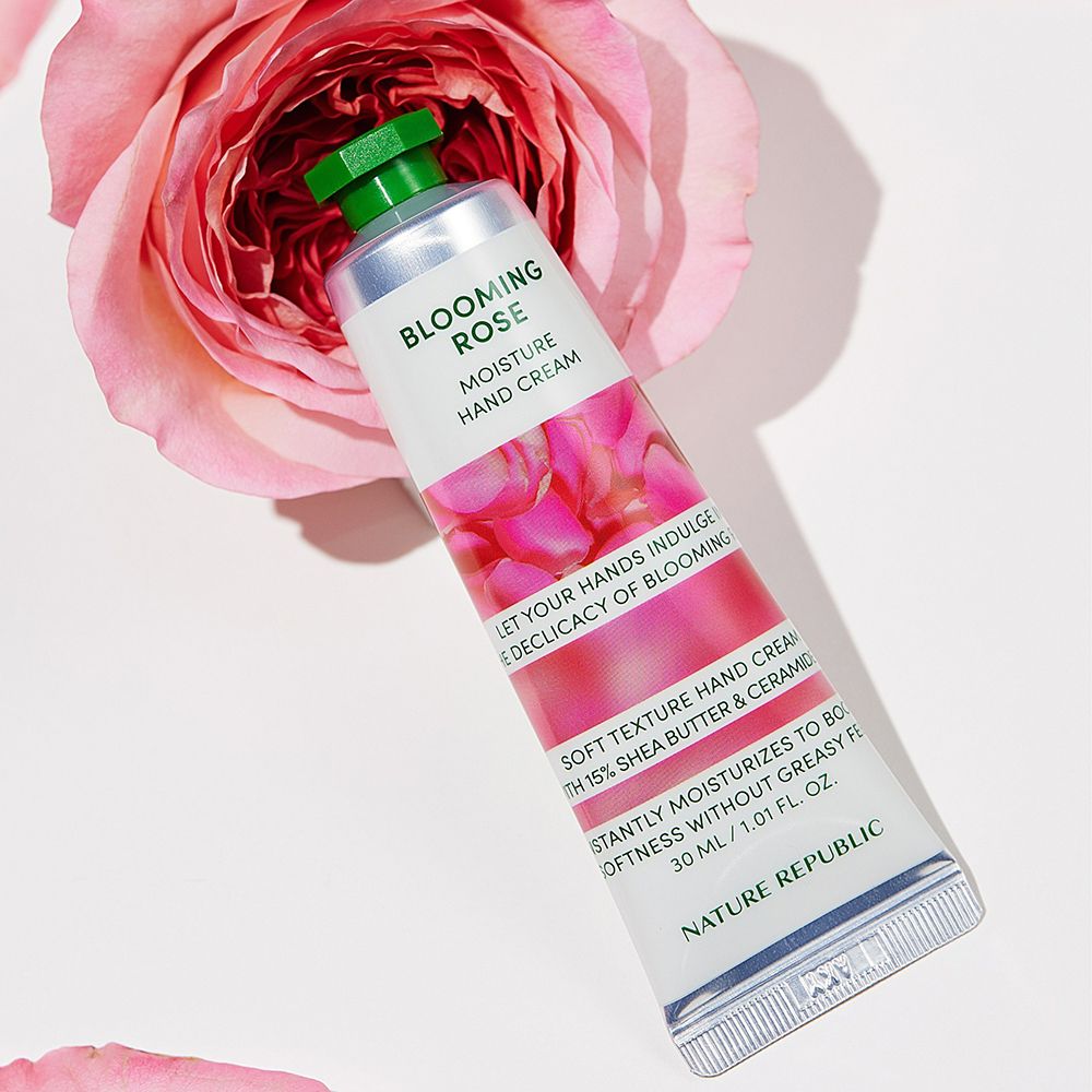 Tube de crème pour les mains, design blanc et rose, bouchon vert. Texte: Blooming Rose, Hand & Nature. Posé à côté d'une rose.