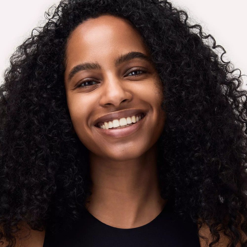 Portrait d'une femme souriante aux cheveux bouclés et foncés. Elle porte un haut noir. Teint naturel, bouche ouverte.