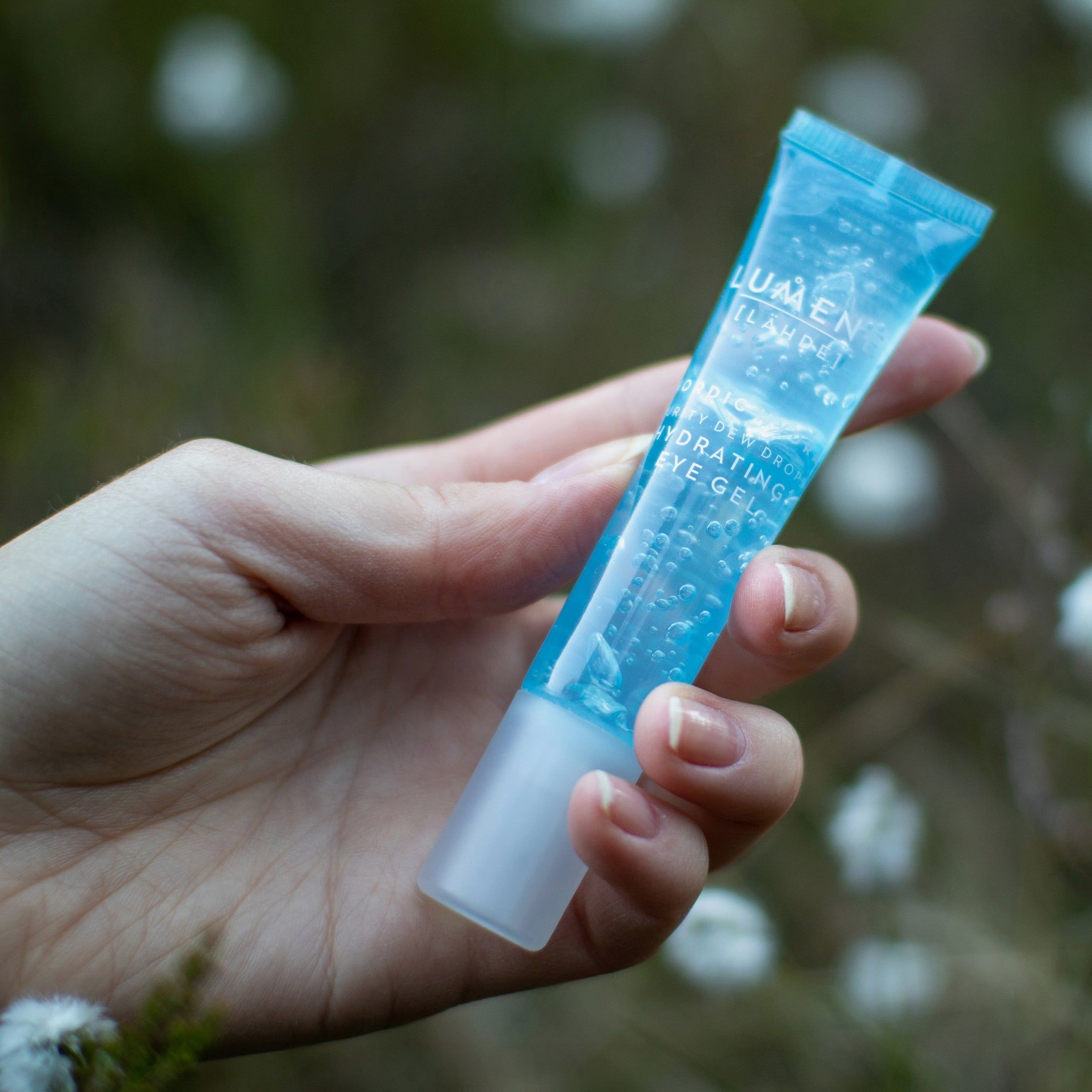 Main tenant un tube bleu. Inscription : Lumene [LAHDE] Nordic Hydra Purity Dew Drops Hydrating Eye Gel. Fond : fleurs floues.