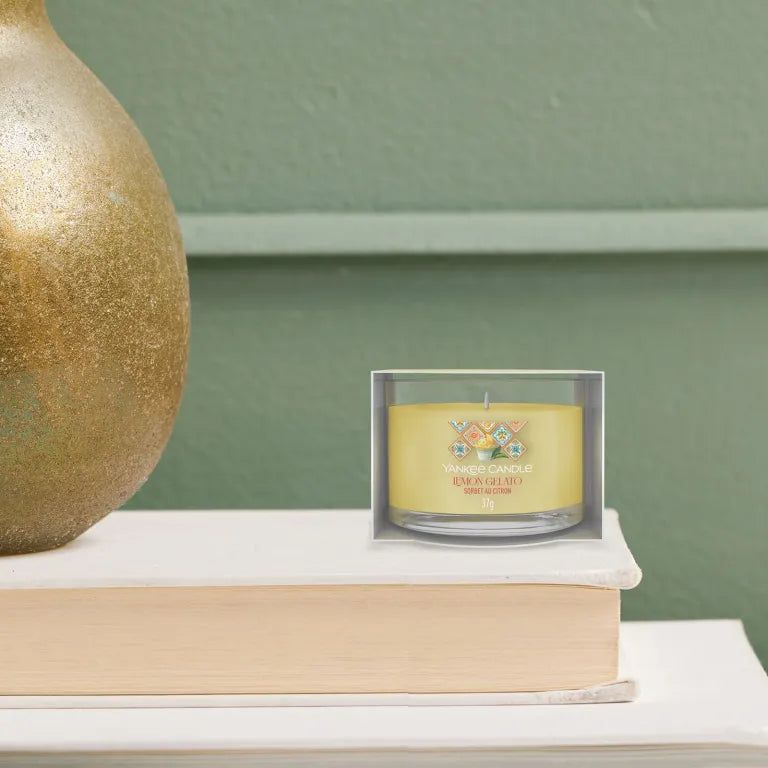 Bougie jaune en verre dans son emballage. Inscription: Lemon Gelato. Posée sur un livre. Fond: mur vert.