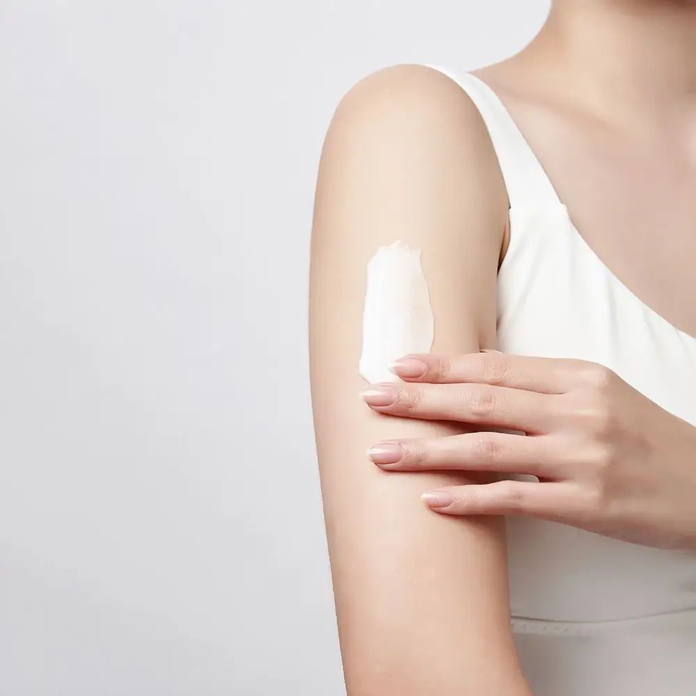 Arm bedekt met witte crème. Persoon draagt een witte top. Lichte achtergrond.