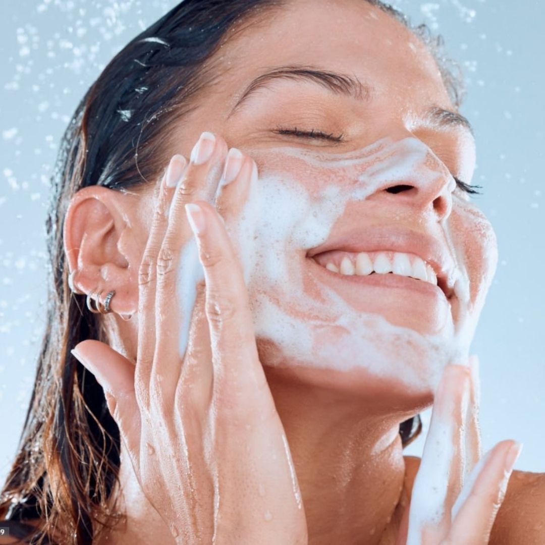 Vrouw wast haar gezicht met witte schuim. Water spettert. Ze glimlacht.