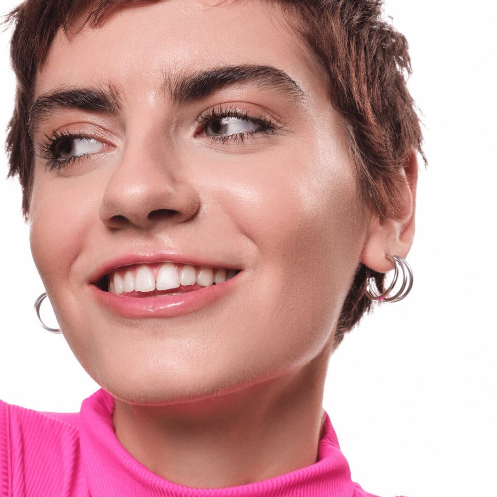 Gros plan d'une femme aux cheveux courts souriant à la caméra. Elle porte des créoles argentées et un haut rose.