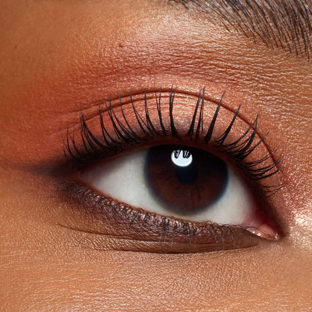 Gros plan sur un œil avec fard à paupières dans les tons marron et orange. Longs cils et sourcils foncés.