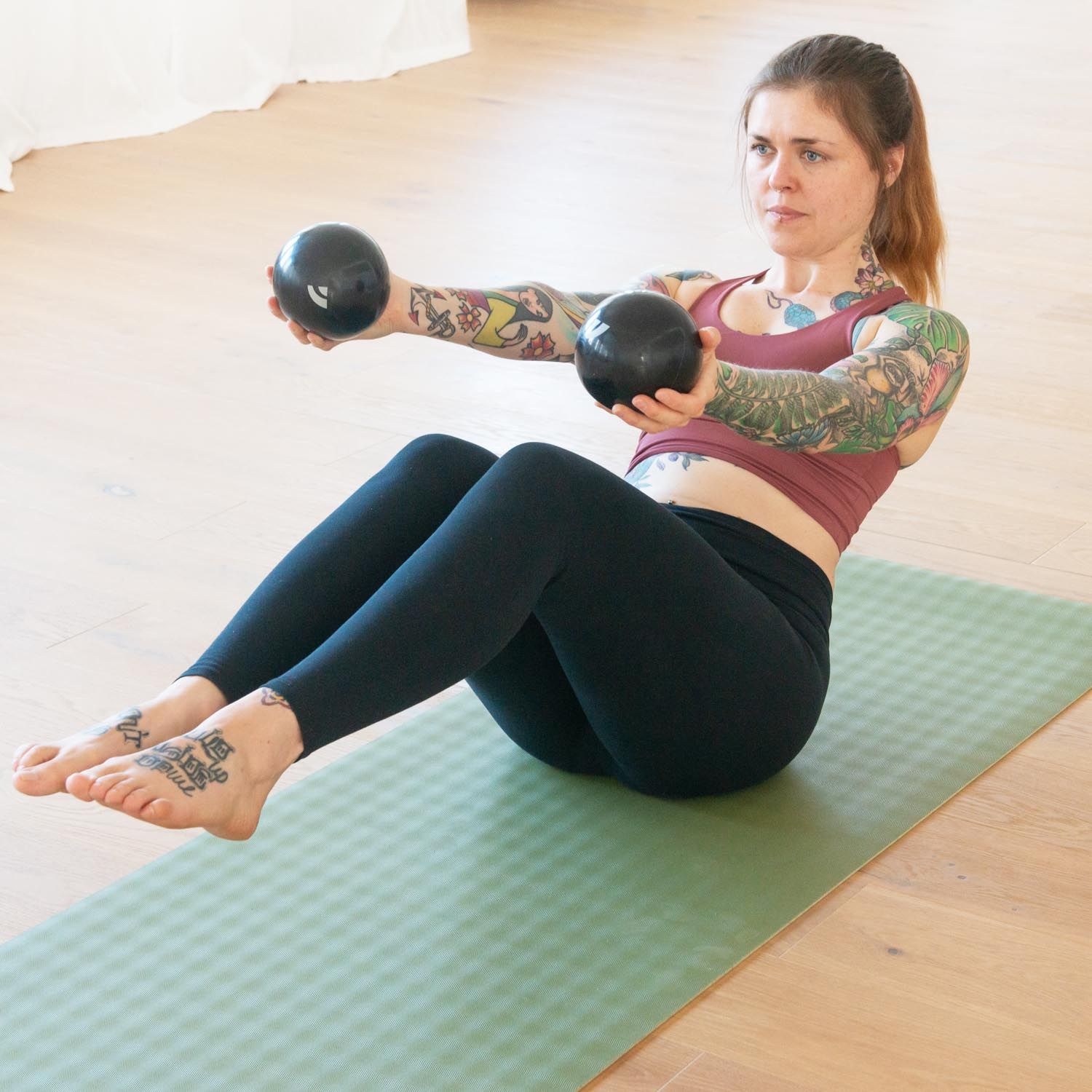 Une femme assise sur un tapis vert tient deux balles de Pilates tonifiantes anthracite. Elle s'entraîne.