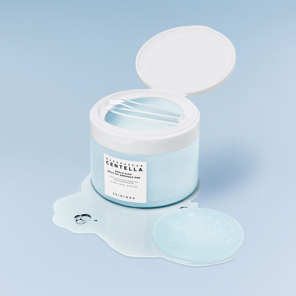 Contenant avec couvercle ouvert, pad et liquide. Texte: CENTELLA, HYALU-CICA JELLY-FIT AMPULE PAD, SKIN1004. Arrière-plan: bleu clair.