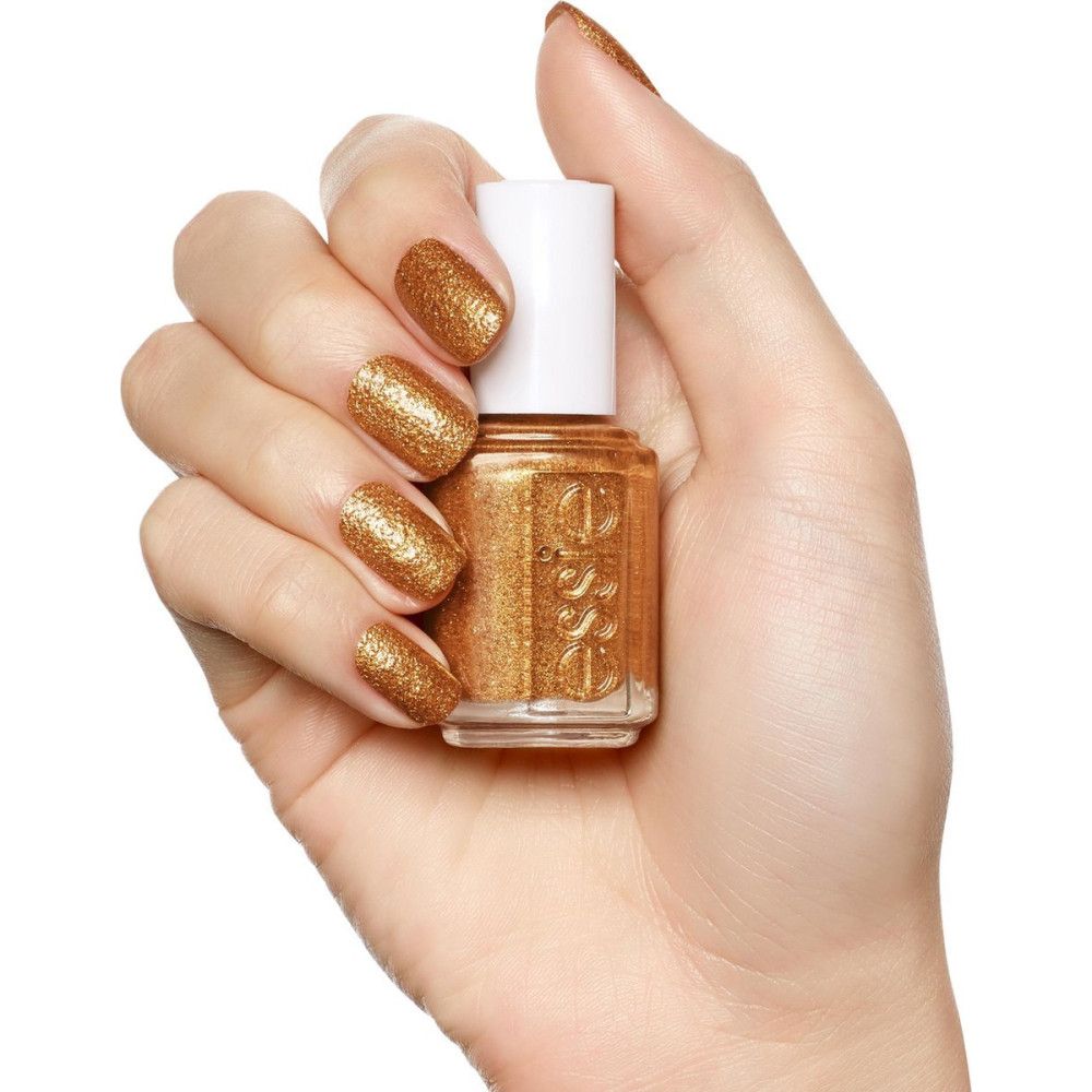 Hand houdt Essie nagellak vast. Gouden lak op nagels en flesje. Witte dop, gouden vloeistof, Essie logo.