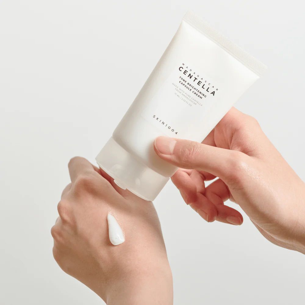 Handen houden een tube vast. Crème wordt op de hand aangebracht. Merk SKIN1004.