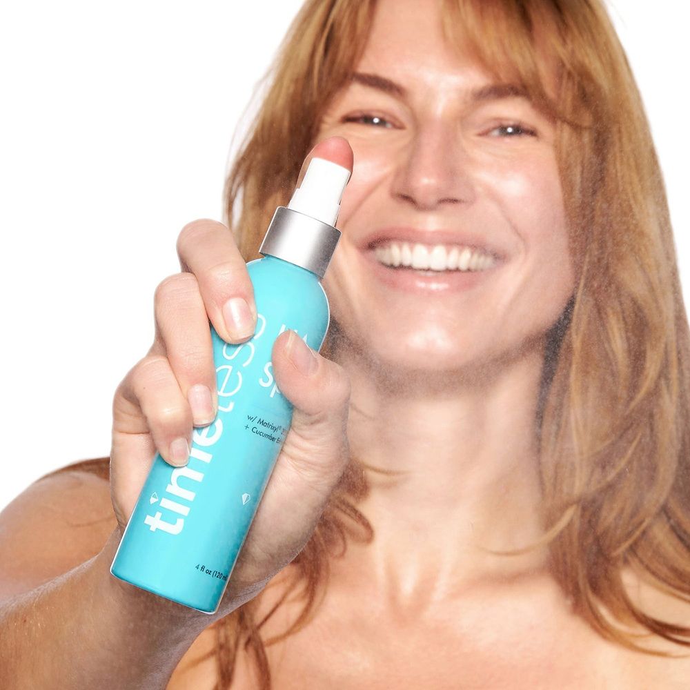 Vrouw spuit met een turquoise fles. Opschrift: timeless, HA Spray, Matrixyl 3000 + Cucumber Extract. Sproeimist zichtbaar.